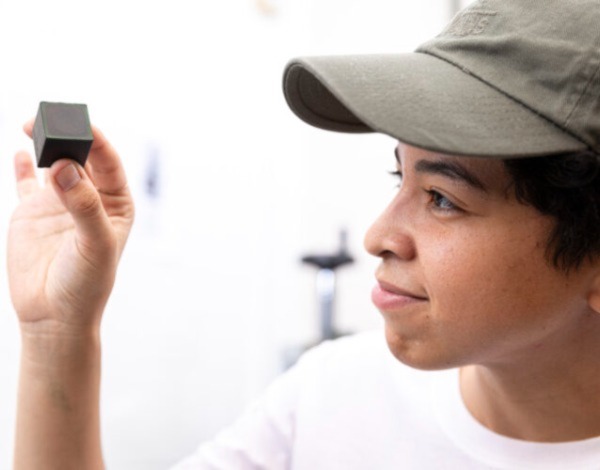 A person wearing a baseball cap holds up a black cube made of bioplastic and is looking at it